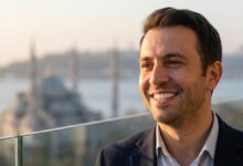 Un homme élégant avec un sourire éclatant et sain, se tenant sur un balcon avec une vue panoramique floue d'Istanbul au lever du soleil en arrière-plan, représentant le succès du tourisme dentaire en Turquie.